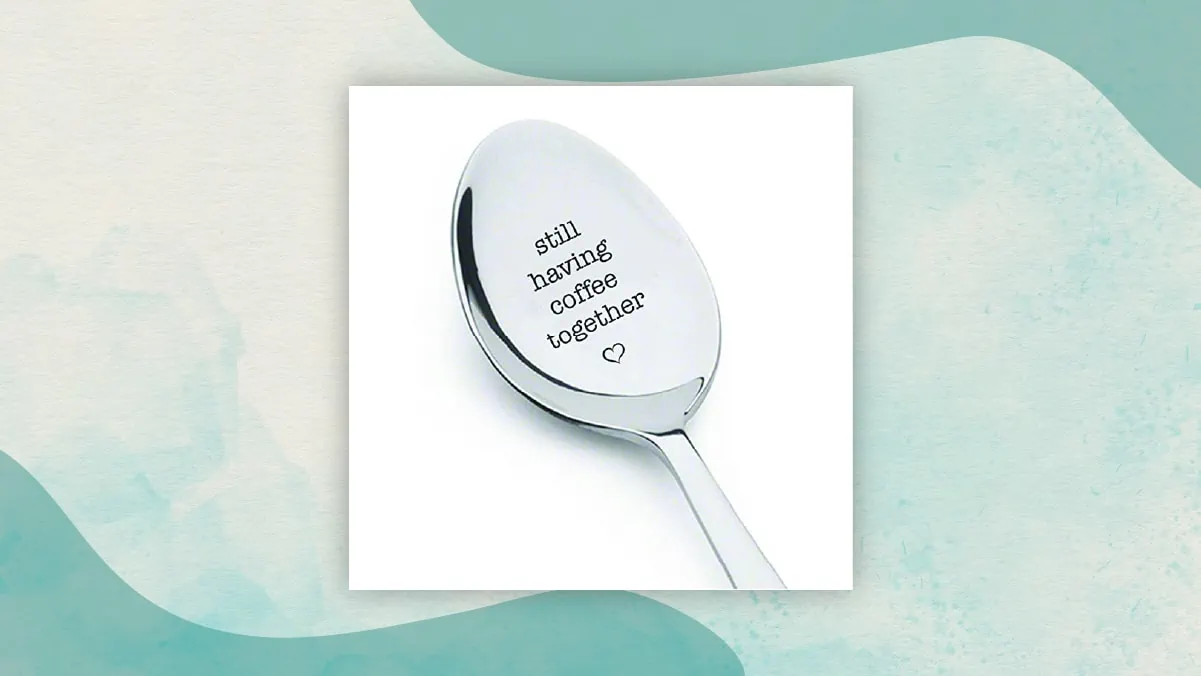 A spoon with "still having coffee together" written on it and a small heart shape below it in a white background. 