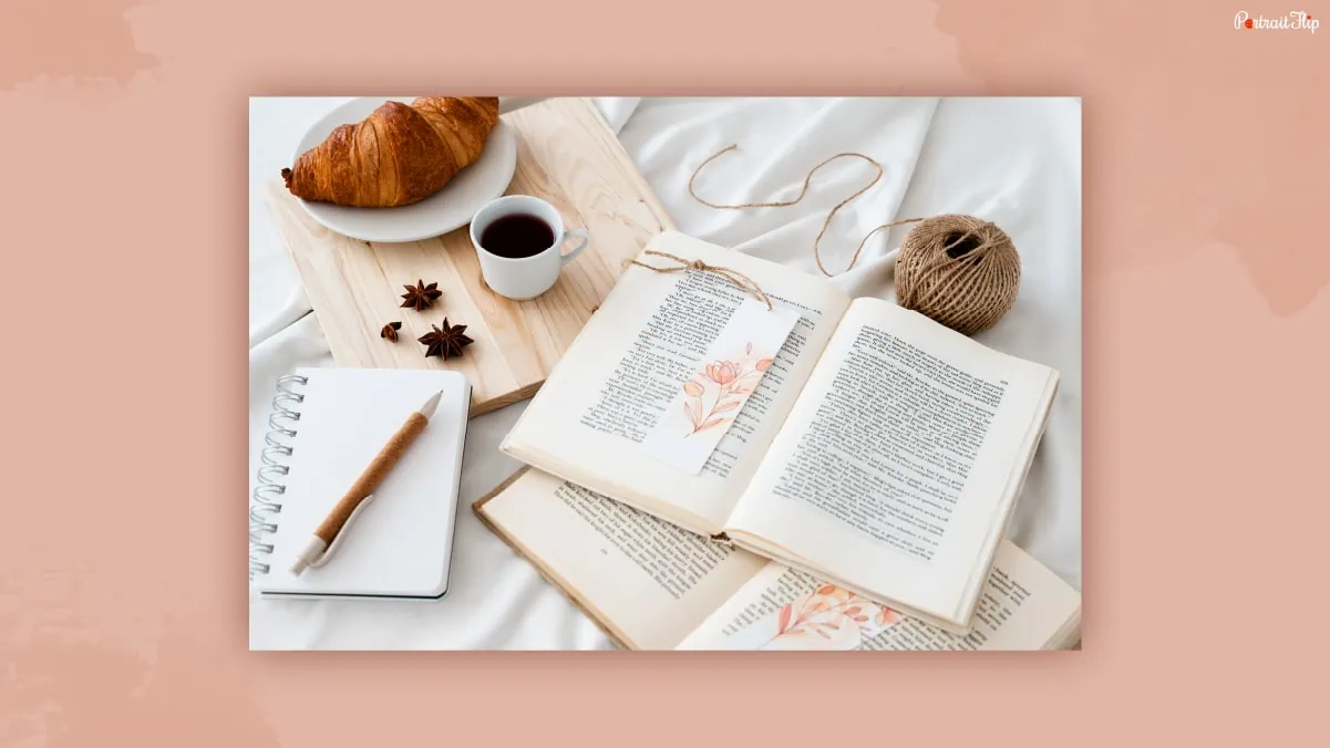 A bookmark kept in between the books beside a blank notebook with croissant, tea and wool on a white bedsheet that could be one of the thank-you gifts for women.